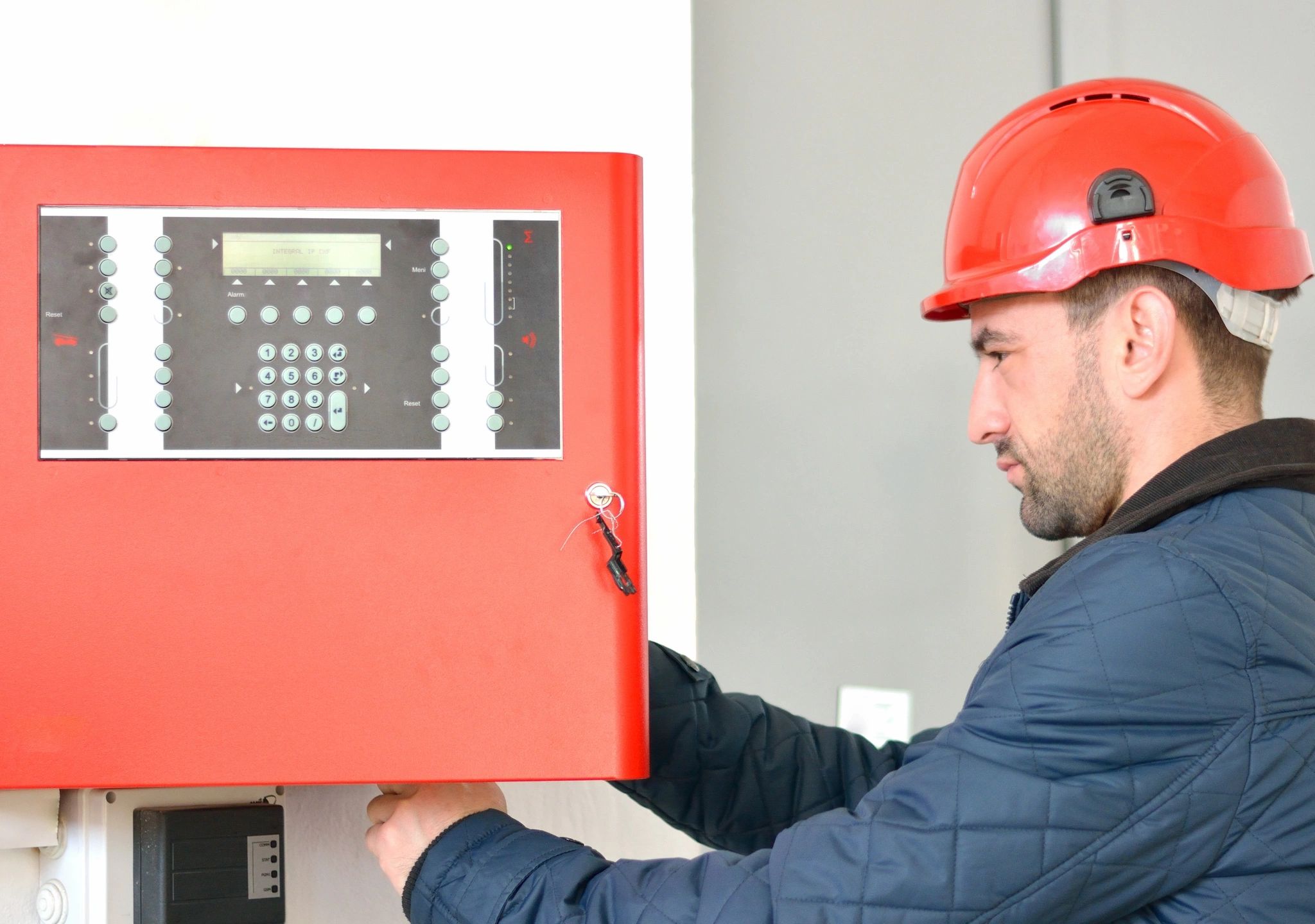 Technician in red hard hat inspecting fire alarm control panel